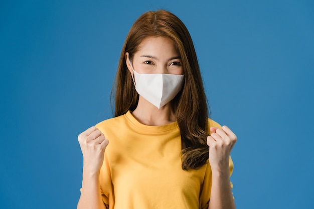 Junges asiatisches Mädchen, das medizinische Gesichtsmaske trägt, die Friedenszeichen zeigt, ermutigt mit gekleidet in lässigem Stoff und Blick auf Kamera lokalisiert auf blauem Hintergrund. Soziale Distanzierung, Quarantäne für Koronavirus.