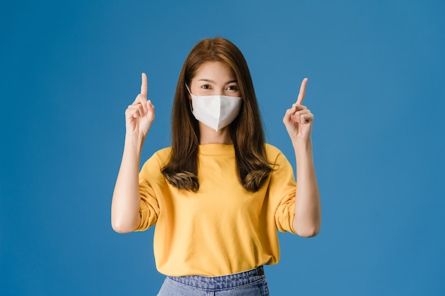 Junges asiatisches Mädchen, das medizinische Gesichtsmaske trägt, zeigt etwas an der leeren Stelle mit gekleidet in lässigem Stoff und betrachtet Kamera lokalisiert auf blauem Hintergrund. Soziale Distanzierung, Quarantäne für Koronavirus.
