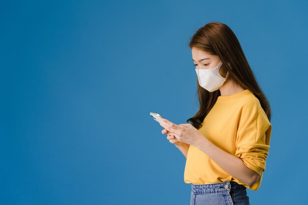 Junges asiatisches Mädchen, das medizinische Gesichtsmaske unter Verwendung des Mobiltelefons mit gekleidet in der Freizeitkleidung lokalisiert auf blauem Hintergrund trägt. Selbstisolation, soziale Distanzierung, Quarantäne zur Vorbeugung von Koronaviren.