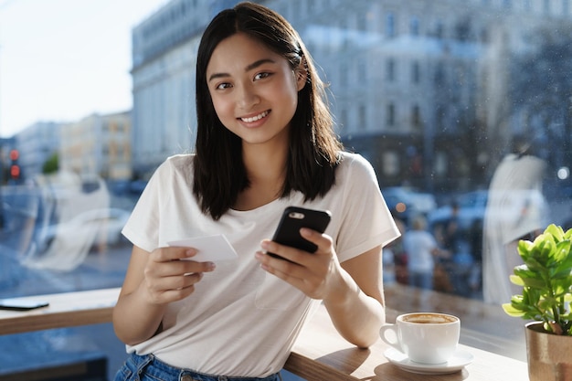 Junges asiatisches mädchen, das mit kreditkarte und smartphone für kaffee bezahlt, lächelt im café nahe dem fenster, das cappuccino trinkt