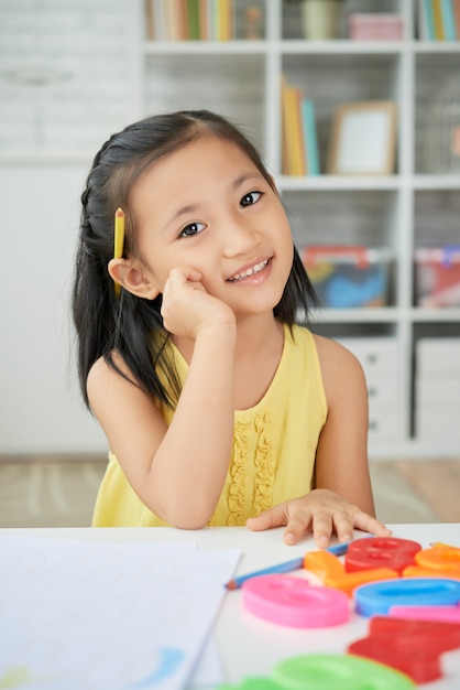 Junges asiatisches Mädchen, das zu Hause, mit der Hand zur Backe, Bleistift hinter Ohr und Plastikzahlen auf Schreibtisch sitzt