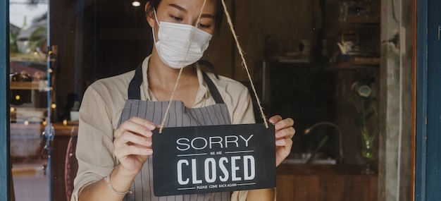 Junges asiatisches Mädchen trägt eine Gesichtsmaske, die nach der Quarantäne der Coronavirus-Sperrung ein Schild vom offenen zum geschlossenen Schild am Glastür-Café umwandelt.