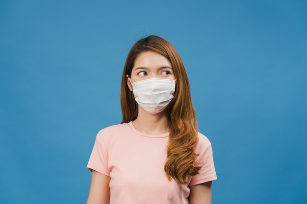Junges asiatisches Mädchen trägt eine medizinische Gesichtsmaske, müde von Stress und Anspannung, schaut selbstbewusst auf den Raum isoliert auf blauer Wand