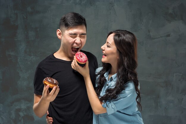 Junges asiatisches Paar genießt das Essen des süßen bunten Donuts auf grauem Studio