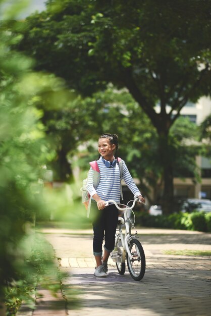 Junges asiatisches Schulmädchen mit Rucksack und Fahrrad gehend durch Park