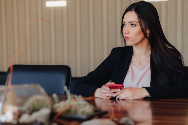 Kostenloses Foto junges attraktives emotionales mädchen in der geschäftsart kleidet das sitzen am schreibtisch mit telefon im büro oder im publikum