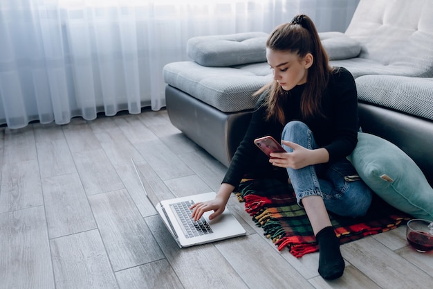 Junges attraktives Mädchen zu Hause, das mit einem Laptop arbeitet und am Telefon spricht. Komfort und Gemütlichkeit zu Hause. Home Office und Arbeit von zu Hause aus. Remote-Online-Beschäftigung.