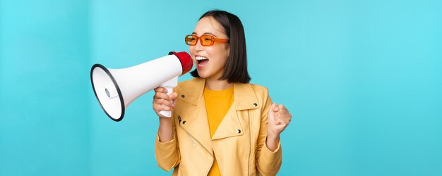 Junges begeistertes koreanisches Mädchen im trendigen Outfit, das im Megaphon schreit und Ankündigungswerbung macht, die in der Freisprecheinrichtung schreit, die über blauem Hintergrund steht