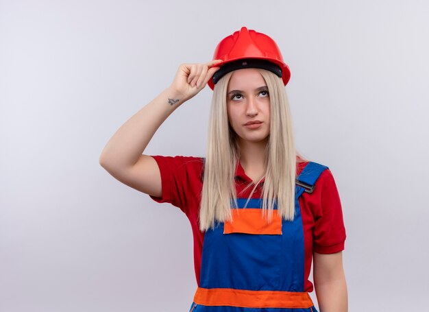 Junges blondes Ingenieur-Baumeistermädchen in der Uniform, die Hand auf Kopf setzt, der auf isolierte weiße Wand schaut