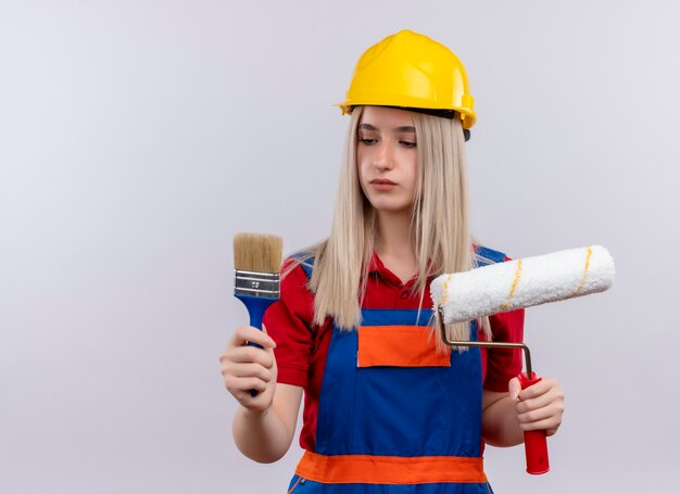 Junges blondes Ingenieur-Baumeistermädchen in der Uniform, die Pinsel und Rolle hält und sie auf isolierter weißer Wand mit Kopienraum betrachtet