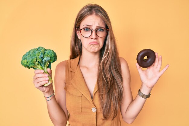 Junges blondes Mädchen, das Brokkoli und Schokoladenkrapfen deprimiert hält und sich Sorgen um Not macht, wütend und ängstlich weint. trauriger Ausdruck.