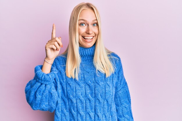 Junges blondes Mädchen, das Wollwinterpullover trägt und mit dem Finger nach oben zeigt, mit erfolgreicher Idee. aufgeregt und glücklich. Nummer Eins.