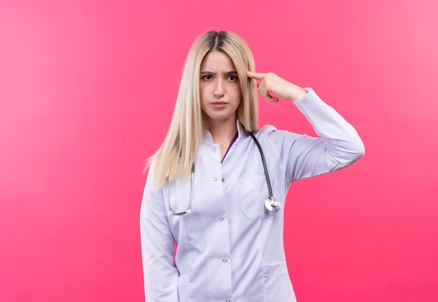 junges blondes Mädchen des Arztes, das Stethoskop im medizinischen Kleid trägt, legte ihren Finger auf Stirn auf isolierte rosa Wand