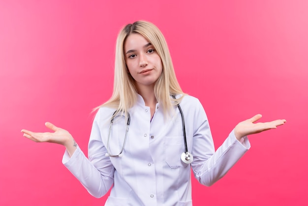 junges blondes Mädchen des Arztes, das Stethoskop im medizinischen Kleid trägt, verbreitet Hände auf isolierter rosa Wand