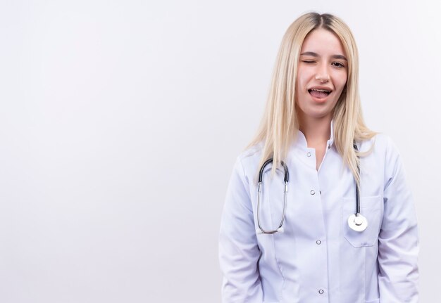 junges blondes Mädchen des blinkenden Doktors, das Stethoskop und medizinisches Kleid in Zahnspange auf isolierter weißer Wand trägt