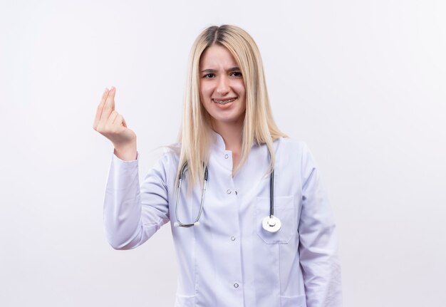 junges blondes Mädchen des Doktors, das Stethoskop und medizinisches Kleid in Zahnspange trägt, die Geldgeste auf isolierter weißer Wand zeigt