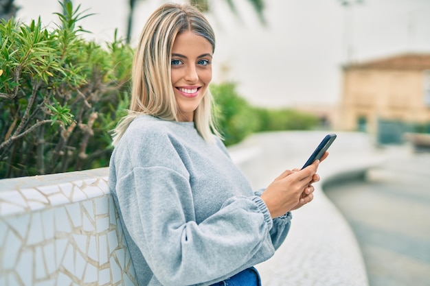 Kostenloses Foto junges blondes mädchen lächelt glücklich und benutzt smartphone, während es auf der bank im park sitzt