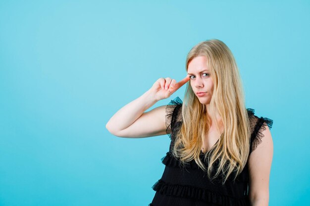 Junges blondes Mädchen schaut in die Kamera, indem sie den Zeigefinger auf der Schläfe auf blauem Hintergrund hält