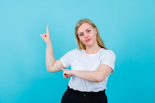 Junges blondes Mädchen zeigt mit dem Zeigefinger auf blauem Hintergrund nach oben