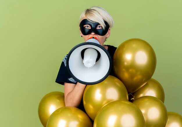 Junges blondes Partei-Mädchen, das Maskerademaske trägt, die hinter Ballons steht, die Kamera betrachten, die durch Sprecher lokalisiert auf olivgrünem Hintergrund mit Kopienraum spricht