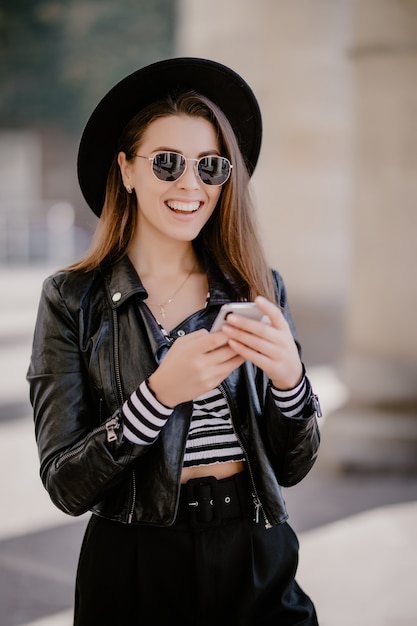 Junges braunhaariges Mädchen in einer Lederjacke, schwarzem Hut auf der Stadtpromenade und spielt auf Handy