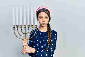 Kostenloses Foto junges brünettes mädchen, das menorah chanukka jüdische kerze hält, die wangen mit lustigem gesicht aufbläht. mund mit luft aufgeblasen, luft fangend.