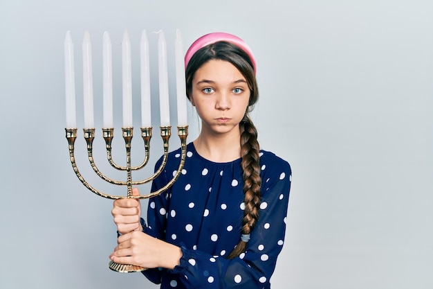 Junges brünettes mädchen, das menorah chanukka jüdische kerze hält, die wangen mit lustigem gesicht aufbläht. Mund mit Luft aufgeblasen, Luft fangend.