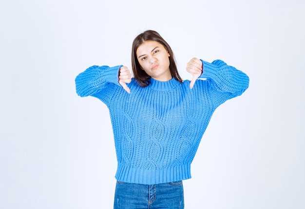 Junges Brunettemädchen in der blauen Strickjacke, die Daumen unten auf Weiß gibt.