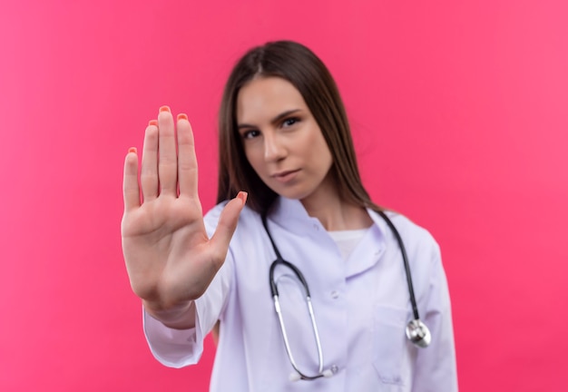 junges Doktormädchen, das medizinisches Stethoskopkleid trägt, das Stoppgeste auf isolierter rosa Wand zeigt