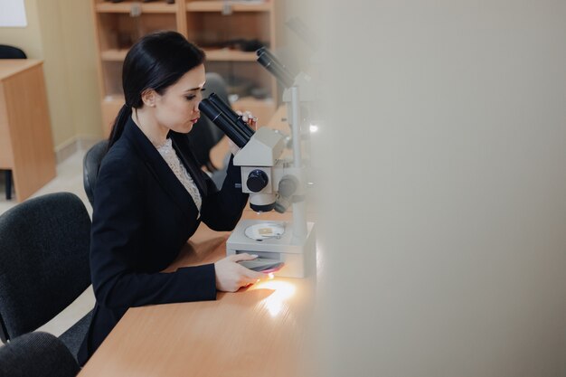 Junges emotionales attraktives Mädchen, das am Tisch sitzt und mit einem Mikroskop in einem modernen Büro oder in einem Publikum arbeitet