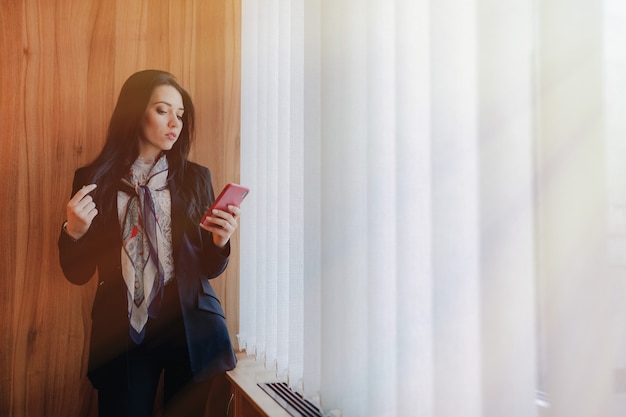 Junges emotionales attraktives Mädchen in der Geschäftsart kleidet an einem Fenster mit einem Telefon in einem modernen Büro oder in einem Auditorium