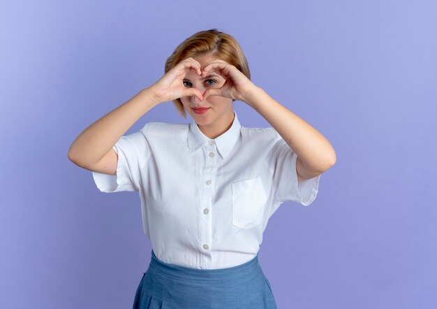 Junges erfreutes blondes russisches Mädchen gestikuliert und schaut durch Herzhandzeichen lokalisiert auf lila Hintergrund mit Kopienraum