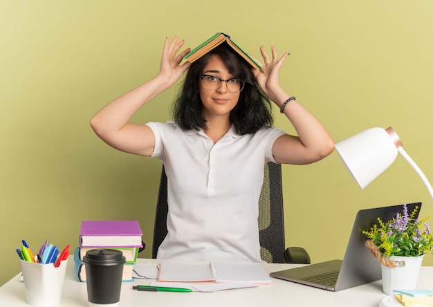 Junges erfreutes hübsches kaukasisches Schulmädchen, das Brille trägt, sitzt am Schreibtisch mit Schulwerkzeugen hält Buch über Kopf auf Grün mit Kopienraum