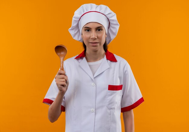Junges erfreutes kaukasisches Kochmädchen in der Kochuniform hält Holzlöffel lokalisiert auf orange Raum mit Kopienraum