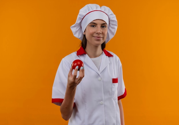 Junges erfreutes kaukasisches Kochmädchen in der Kochuniform hält Tomate, die Kamera auf Orange mit Kopienraum betrachtet