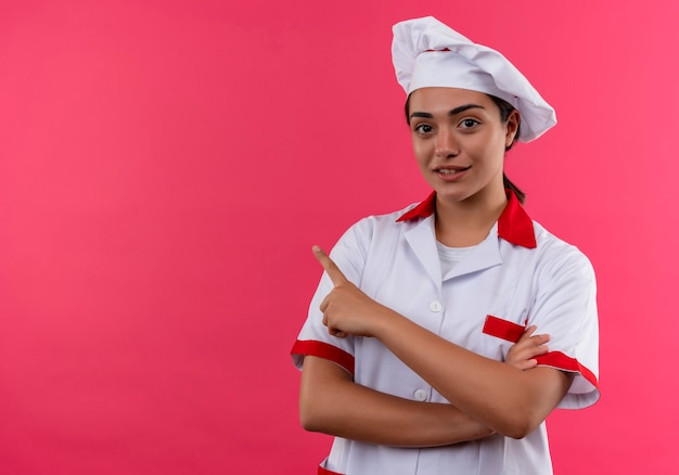 Junges erfreutes kaukasisches Kochmädchen in der Kochuniform kreuzt Arme und zeigt auf die Seite, die auf rosa Wand mit Kopienraum isoliert ist