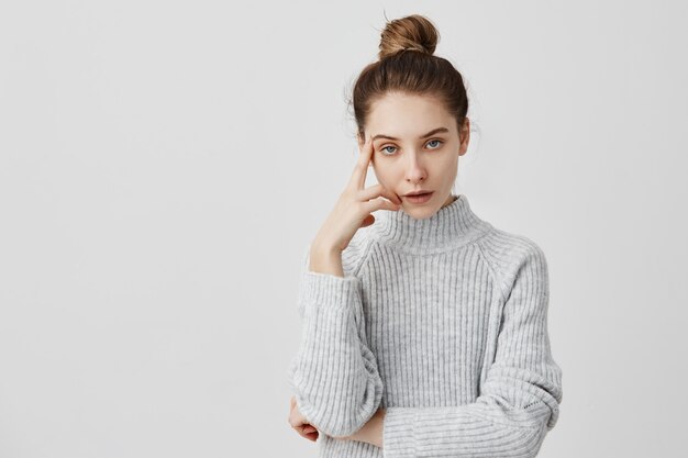Junges erwachsenes Mädchen posiert mit genervtem Blick, der Finger auf Schläfe hält. Attraktive Frau im grauen Kleid müde vom Hören uninteressanter Geschichten. Kommunikationskonzept