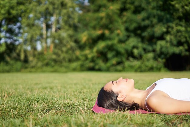 Junges Fitness-Mädchen, das auf einer Sportmatte auf dem Rasen liegt und im Park in Sportkleidung atmet und meditiert