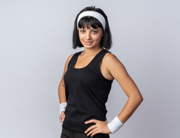 Junges Fitness-Mädchen mit Stirnband, das die Kamera anschaut und selbstbewusst stehend über weiß lächelt