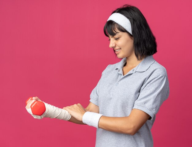 Junges Fitness-Mädchen mit Stirnband, das eine Hantel in ihrer bandagierten Hand hält und sich Unbehagen und Schmerzen über rosa stehend fühlt