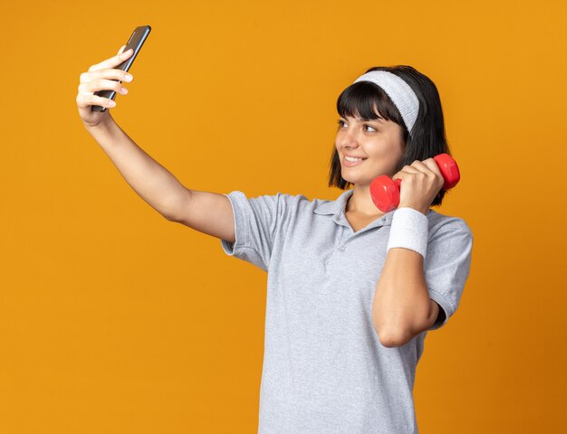 Junges Fitness-Mädchen mit Stirnband, das Hantel hält, macht Selfie mit Smartphone und lächelt fröhlich über Orange stehend
