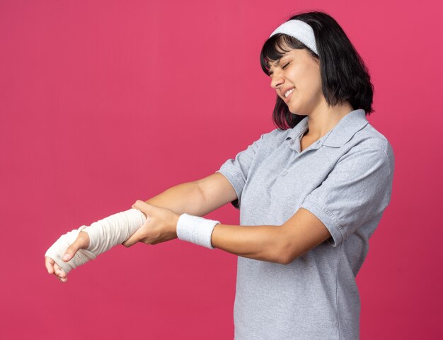 Junges Fitness-Mädchen mit Stirnband, das ihre bandagierte Hand berührt, sieht unwohl aus und fühlt Schmerzen auf rosafarbenem Hintergrund
