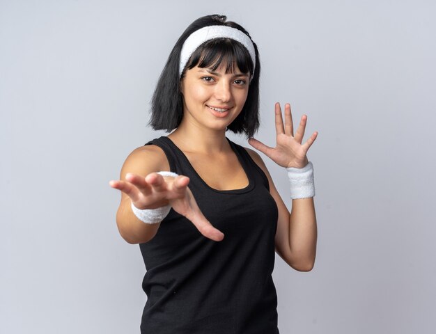Junges Fitness-Mädchen mit Stirnband, das in die Kamera schaut und mit den Händen gestikuliert, die fröhlich lächeln, die über weißem Hintergrund stehen