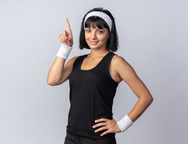 Junges Fitness-Mädchen mit Stirnband, das in die Kamera schaut und selbstbewusst mit dem Zeigefinger zeigt, der über Weiß steht