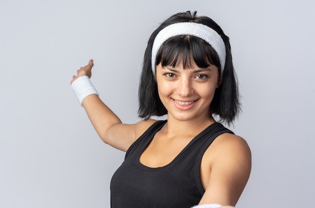 Junges Fitness-Mädchen mit Stirnband, das mit einem Lächeln auf einem glücklichen Gesicht auf weißem Hintergrund in die Kamera schaut