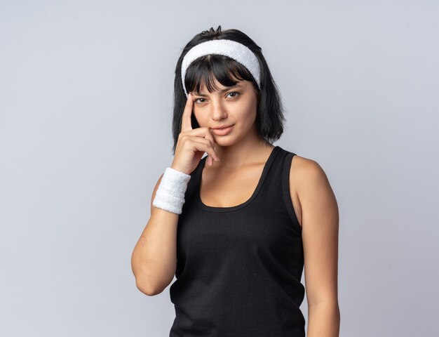 Junges Fitness-Mädchen mit Stirnband, das mit einem Lächeln auf einem intelligenten Gesicht in die Kamera schaut und mit dem Zeigefinger auf ihre Schläfe zeigt, die über weißem Hintergrund steht