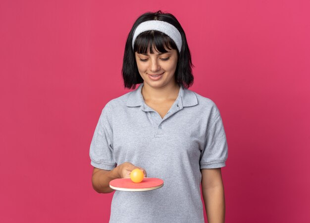 Junges Fitness-Mädchen mit Stirnband, das Schläger und Ball für Tischtennis hält und mit glücklichem Gesicht auf rosafarbenem Hintergrund lächelt