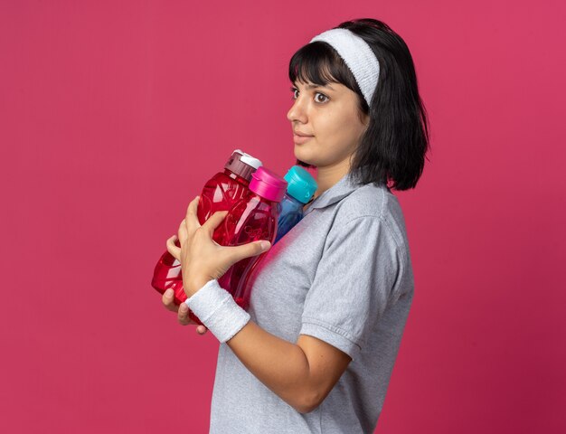 Junges Fitness-Mädchen mit Stirnband, das Wasserflaschen hält und verwirrt beiseite schaut, steht auf rosa Hintergrund