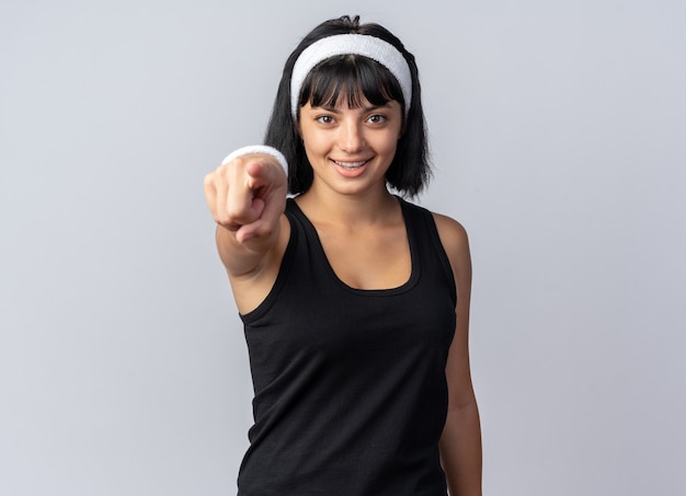 Junges Fitness-Mädchen mit Stirnband glücklich und selbstbewusst, das mit dem Zeigefinger auf die Kamera zeigt, die über weißem Hintergrund steht