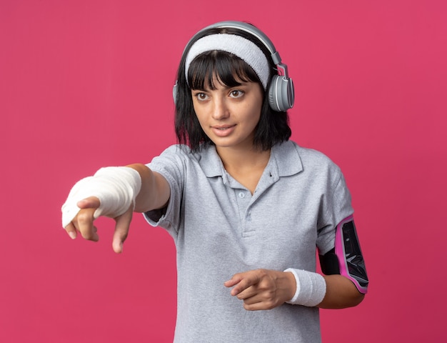 Junges Fitness-Mädchen mit Stirnband mit Kopfhörern und Armband für Smartphone, das beiseite schaut und mit dem Zeigefinger auf etwas zeigt, das auf rosafarbenem Hintergrund steht